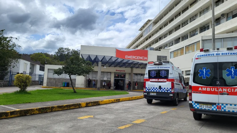 na madre angustiada con su hijo pequeño en brazos, en el exterior de un hospital en Cuenca, con una ambulancia de fondo, ilustrando la crisis sanitaria y el pago por un servicio vital.